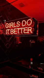 A neon sign with pink and red colors illuminating the words 'Girls Do It Better' against a dark tiled background. The glow of the neon lights casts a warm, vibrant hue on the surrounding surfaces.