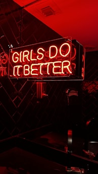 A neon sign with pink and red colors illuminating the words 'Girls Do It Better' against a dark tiled background. The glow of the neon lights casts a warm, vibrant hue on the surrounding surfaces.