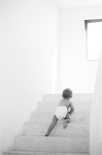 A young child in a diaper is crawling up a set of white stairs. The environment is minimalistic, with bright natural light coming from a window at the top right corner.