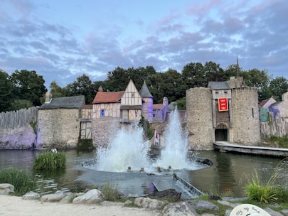 A medieval-style fortress with stone walls, a wooden gate, and timber-framed buildings. A moat surrounds the structure, with water fountains creating large sprays in the center. The setting is lush with green trees in the background and a partially cloudy sky.
