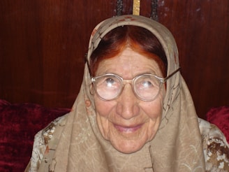 An elderly woman with glasses smiles warmly. She wears a patterned headscarf and has visible wrinkles on her face, reflecting age and experience. The background is a dark wood panel, and part of a red cushion or blanket is visible at the bottom.