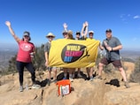 A group of seven people poses on a rocky hilltop holding a yellow banner with the words 'World Cleanup Day.' They are dressed in casual outdoor clothing, and several have hats on. Around them is a clear blue sky and a distant view of a cityscape and hills.