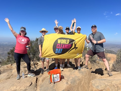 A group of seven people poses on a rocky hilltop holding a yellow banner with the words 'World Cleanup Day.' They are dressed in casual outdoor clothing, and several have hats on. Around them is a clear blue sky and a distant view of a cityscape and hills.