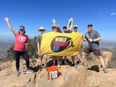 A group of seven people poses on a rocky hilltop holding a yellow banner with the words 'World Cleanup Day.' They are dressed in casual outdoor clothing, and several have hats on. Around them is a clear blue sky and a distant view of a cityscape and hills.
