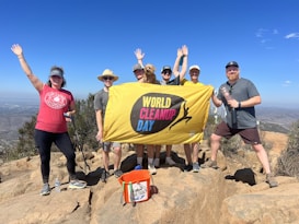A group of seven people poses on a rocky hilltop holding a yellow banner with the words 'World Cleanup Day.' They are dressed in casual outdoor clothing, and several have hats on. Around them is a clear blue sky and a distant view of a cityscape and hills.