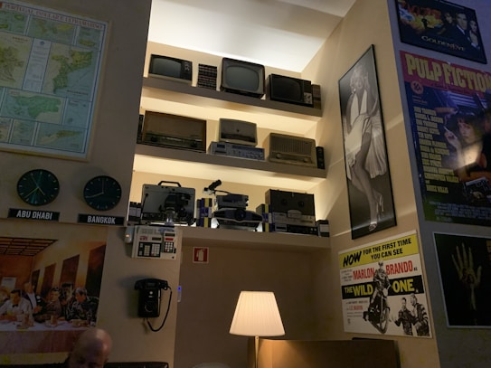 The interior features an eclectic arrangement of vintage technology and pop culture elements. Multiple old televisions, radios, and film projectors are neatly displayed on shelves. A black rotary wall phone and a table lamp are situated below, alongside retro wall clocks labeled 'Abu Dhabi' and 'Bangkok.' The walls are adorned with classic movie posters including 'Pulp Fiction,' and a famous black and white portrait.