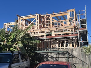A multi-story building under construction with a wooden framework. Scaffolding surrounds the structure, and there are green leafy plants to the left and vehicles parked at the front. The sky is clear and blue.