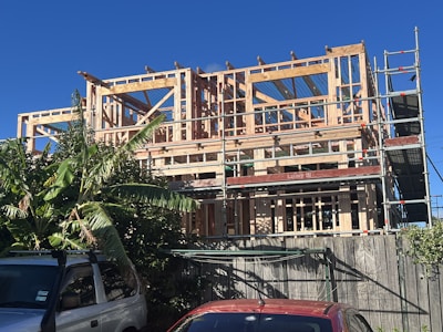 A multi-story building under construction with a wooden framework. Scaffolding surrounds the structure, and there are green leafy plants to the left and vehicles parked at the front. The sky is clear and blue.