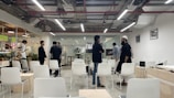 A modern office space with a group of people engaged in conversation. The room has white chairs and tables, some occupied by people, and is well-lit with overhead lights. There are visible plants and glass walls with sticky notes in the background.