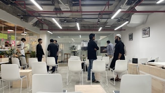A modern office space with a group of people engaged in conversation. The room has white chairs and tables, some occupied by people, and is well-lit with overhead lights. There are visible plants and glass walls with sticky notes in the background.