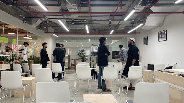 A modern office space with a group of people engaged in conversation. The room has white chairs and tables, some occupied by people, and is well-lit with overhead lights. There are visible plants and glass walls with sticky notes in the background.