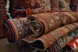 Several intricately designed, colorful rugs are rolled up and stacked on top of each other. The patterns are detailed, featuring geometric shapes and floral motifs, predominantly in shades of red, blue, and brown.