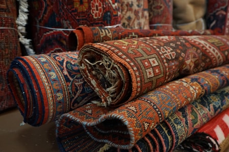 Several intricately designed, colorful rugs are rolled up and stacked on top of each other. The patterns are detailed, featuring geometric shapes and floral motifs, predominantly in shades of red, blue, and brown.