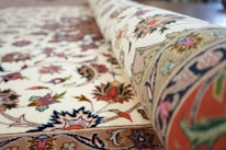 A close-up of a rolled, intricately patterned carpet with floral and geometric designs. The carpet displays a mix of colors including reds, blues, greens, and pinks against a cream background. The detailed embroidery and craftsmanship are evident in the texture and vibrant patterns.