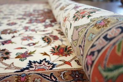 A close-up of a rolled, intricately patterned carpet with floral and geometric designs. The carpet displays a mix of colors including reds, blues, greens, and pinks against a cream background. The detailed embroidery and craftsmanship are evident in the texture and vibrant patterns.