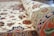 A close-up of a rolled, intricately patterned carpet with floral and geometric designs. The carpet displays a mix of colors including reds, blues, greens, and pinks against a cream background. The detailed embroidery and craftsmanship are evident in the texture and vibrant patterns.