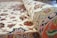 A close-up of a rolled, intricately patterned carpet with floral and geometric designs. The carpet displays a mix of colors including reds, blues, greens, and pinks against a cream background. The detailed embroidery and craftsmanship are evident in the texture and vibrant patterns.