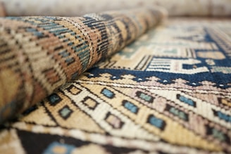 A close-up view of a textured woven rug with intricate geometric patterns in various colors. The rug is partially rolled, enhancing the visibility of its detailed design and structure.