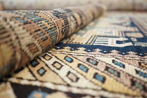 A close-up view of a textured woven rug with intricate geometric patterns in various colors. The rug is partially rolled, enhancing the visibility of its detailed design and structure.