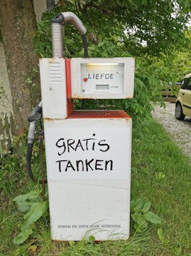 An old-fashioned gas pump with the words 'LIEFDE' and 'GRATIS TANKEN' written on it. The pump is placed outdoors surrounded by greenery, and there is a yellow car partially visible in the background.