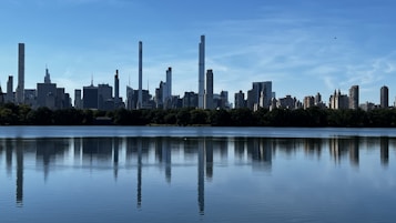 A clear, calm lake reflects the tall skyscrapers silhouetted against a bright blue sky. The skyline features a variety of architectural styles and heights, with modern skyscrapers dominating the scene. The water provides a mirror-like surface that beautifully captures the skyline.