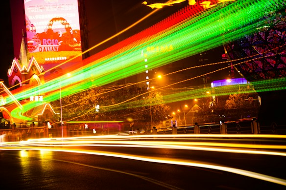 Bright neon light trails in various colors, primarily green and yellow, sweep through an urban night scene. A large illuminated building and streetlights add to the vibrant atmosphere, creating a dynamic and energetic visual effect.