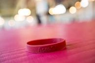 A red silicone wristband is lying on a pink textured surface. The background features soft, out-of-focus lights creating a warm ambiance.