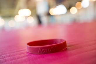 A red silicone wristband is lying on a pink textured surface. The background features soft, out-of-focus lights creating a warm ambiance.