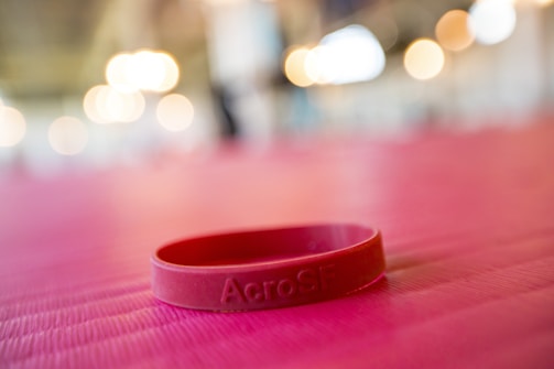 A red silicone wristband is lying on a pink textured surface. The background features soft, out-of-focus lights creating a warm ambiance.