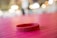 A red silicone wristband is lying on a pink textured surface. The background features soft, out-of-focus lights creating a warm ambiance.