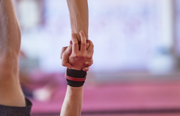Two individuals are engaged in hand balancing, with one person supporting the other by the wrist. The focus is on the hands, which are clasped securely, with skin and fitness straps visible. The background is blurred, suggesting an indoor setting.