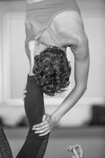 A person is performing a challenging yoga pose that involves an intense backbend and leg stretch. The individual is holding their leg behind their head while standing on one leg, displaying significant flexibility and balance. The background is blurred, making the person the focal point of the image.