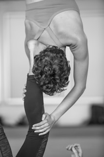 A person is performing a challenging yoga pose that involves an intense backbend and leg stretch. The individual is holding their leg behind their head while standing on one leg, displaying significant flexibility and balance. The background is blurred, making the person the focal point of the image.