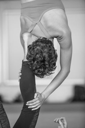 A person is performing a challenging yoga pose that involves an intense backbend and leg stretch. The individual is holding their leg behind their head while standing on one leg, displaying significant flexibility and balance. The background is blurred, making the person the focal point of the image.