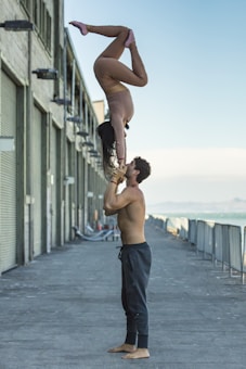 A man stands shirtless, balancing a woman upside down on his hands. They are positioned outdoors, next to a row of industrial buildings with closed roll-up doors. The woman is dressed in a tight, beige outfit, while the man wears dark sweatpants. The background shows a distant view of the ocean and a clear sky.