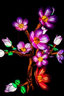 A vibrant display of illuminated flowers with petals in shades of purple, pink, and red, and centers that are yellow. The flowers are attached to stems that appear to be designed in an artistic, whimsical manner. The image is set against a dark background, enhancing the glowing effect of the flowers.