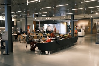 A spacious cafe area with modern decor, featuring people sitting at tables and couches, engaging in conversation and reading. The space is well-lit with overhead lights and has displays of art and artifacts. Wooden chairs and glass display cases add to the ambiance.