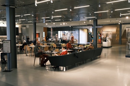 A spacious cafe area with modern decor, featuring people sitting at tables and couches, engaging in conversation and reading. The space is well-lit with overhead lights and has displays of art and artifacts. Wooden chairs and glass display cases add to the ambiance.