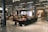 A spacious cafe area with modern decor, featuring people sitting at tables and couches, engaging in conversation and reading. The space is well-lit with overhead lights and has displays of art and artifacts. Wooden chairs and glass display cases add to the ambiance.