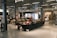 A spacious cafe area with modern decor, featuring people sitting at tables and couches, engaging in conversation and reading. The space is well-lit with overhead lights and has displays of art and artifacts. Wooden chairs and glass display cases add to the ambiance.