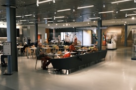 A spacious cafe area with modern decor, featuring people sitting at tables and couches, engaging in conversation and reading. The space is well-lit with overhead lights and has displays of art and artifacts. Wooden chairs and glass display cases add to the ambiance.