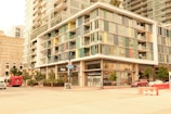 A modern urban scene featuring a multistory building with colorful paneling and large glass windows. The structure includes retail shops on the ground floor and residential levels above. The street is lined with trees, and there are vehicles, including a red bus and a car, as well as construction barriers on the road. Street signs and traffic lights are visible, indicating a busy city intersection.
