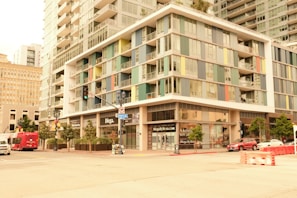 A modern urban scene featuring a multistory building with colorful paneling and large glass windows. The structure includes retail shops on the ground floor and residential levels above. The street is lined with trees, and there are vehicles, including a red bus and a car, as well as construction barriers on the road. Street signs and traffic lights are visible, indicating a busy city intersection.