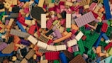 A scattered collection of multicolored LEGO bricks in various shapes and sizes covers the entire space. The blocks include a diverse palette of colors such as red, blue, green, yellow, orange, and white, providing a vibrant and chaotic appearance. Some bricks are rectangular, while others are more unique in shape, forming a jumbled mosaic.