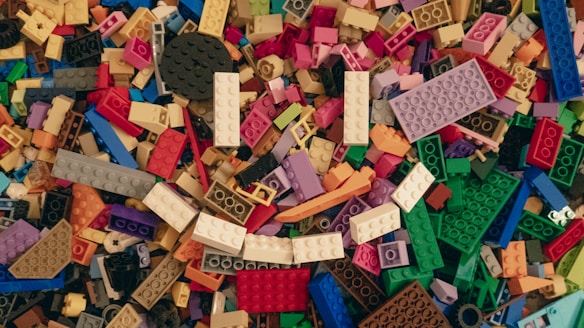 A scattered collection of multicolored LEGO bricks in various shapes and sizes covers the entire space. The blocks include a diverse palette of colors such as red, blue, green, yellow, orange, and white, providing a vibrant and chaotic appearance. Some bricks are rectangular, while others are more unique in shape, forming a jumbled mosaic.