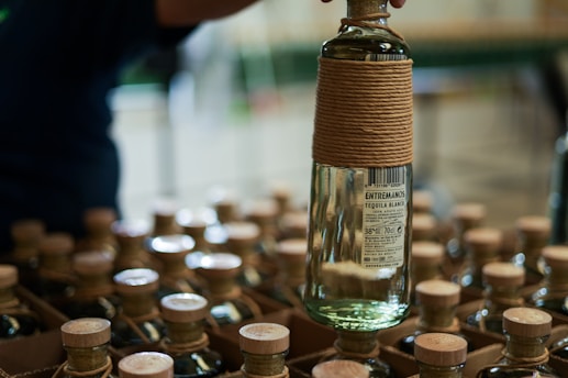 A person holds a bottle of tequila wrapped with twine, amidst a collection of similar bottles neatly organized in boxes. The label reads 'Entremanos Tequila Blanco'. The arrangement suggests an organized setting, likely a store or storage room.
