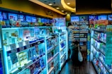 The scene shows a brightly lit drugstore or pharmacy interior with aisles filled with a wide variety of products, including skincare, healthcare items, and everyday essentials. There are informative and promotional signs in multiple languages, mostly focused on East Asian products. Two people are browsing the aisles, one wearing a puffy jacket and the other crouching near the shelves.