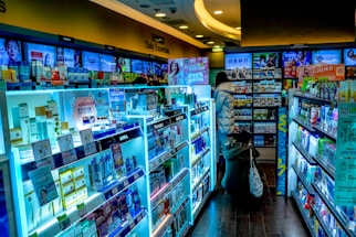 The scene shows a brightly lit drugstore or pharmacy interior with aisles filled with a wide variety of products, including skincare, healthcare items, and everyday essentials. There are informative and promotional signs in multiple languages, mostly focused on East Asian products. Two people are browsing the aisles, one wearing a puffy jacket and the other crouching near the shelves.