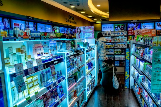 The scene shows a brightly lit drugstore or pharmacy interior with aisles filled with a wide variety of products, including skincare, healthcare items, and everyday essentials. There are informative and promotional signs in multiple languages, mostly focused on East Asian products. Two people are browsing the aisles, one wearing a puffy jacket and the other crouching near the shelves.