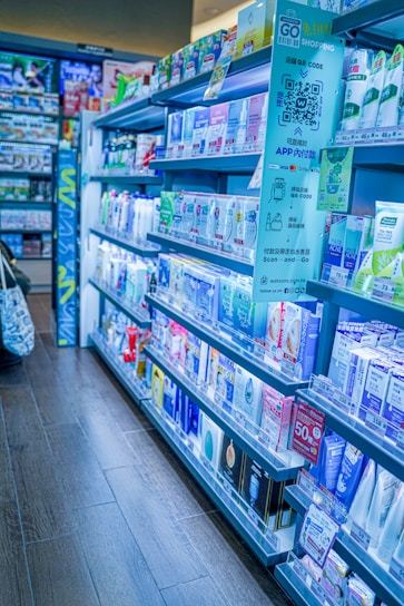 Shelves filled with a variety of skincare and personal care products inside a well-organized pharmacy or store. The shelves are stocked with creams, lotions, and other health-related items, with labels and signs displaying prices and promotions. The aisle has a clean, modern appearance with clear signage for a shopping app.